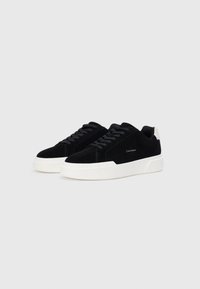 Adidași din suede negru cu o talpă din cauciuc albă, având vârful rotunjit, sistem de închidere cu șireturi și un logo mic pe panoul lateral.