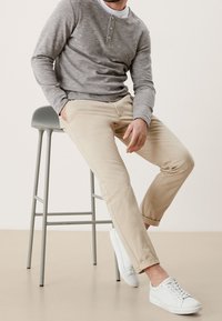 Homme assis sur un tabouret de bar gris, portant un pantalon beige, des baskets blanches, un henley gris à manches longues et un sous-vêtement blanc.
