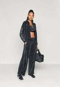 Conjunto de veludo preto com casaco com fecho éclair e calças largas. O traje apresenta uma textura subtil e um ajuste atlético, combinado com ténis.