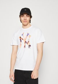 Camiseta de algodón blanca con un gran logotipo de NY colorido en estilo acuarela. Combinada con una gorra negra que tiene un logotipo blanco.