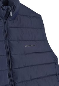 Granatowa, pikowana kamizelka z pionowym przeszyciem, wysokim kołnierzem i zamkiem błyskawicznym z przodu. Posiada srebrne logo po lewej stronie. Miękka tekstura.