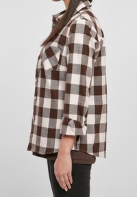 Camicia di flanella a quadri marrone e bianca con maniche arrotolate, due tasche frontali e bottoni rosa, caratterizzata da una vestibilità comoda e da una texture morbida.