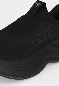 Juodi sportiniai bateliai slip-on tipo su tekstūruotu mezginiu viršuje, su prekės ženklu „HOKA“ šone ir storu paminkštintu padu bėgimui arba vaikščiojimui.