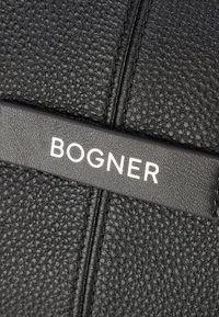 Cuir noir texturé avec grain visible, présentant une sangle noire embossée avec "BOGNER" en blanc. Design épuré, matériel minimal.