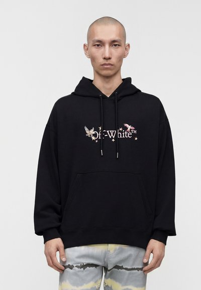 Jeune homme à la tête rasée portant un sweat à capuche noir avec un motif d’oiseau et d’étoile et un pantalon clair tie-dye, debout devant un fond clair uni.