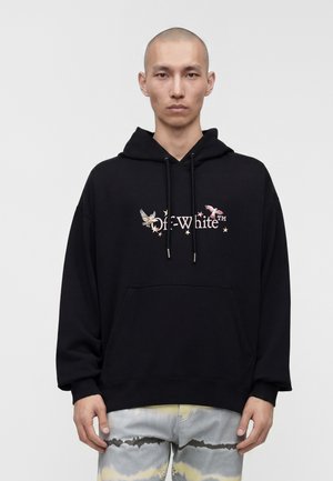 Jeune homme à la tête rasée portant un sweat à capuche noir avec un motif d’oiseau et d’étoile et un pantalon clair tie-dye, debout devant un fond clair uni.