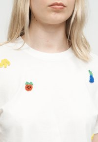 Gros plan sur une personne portant une chemise blanche ornée de petits motifs de fruits perlés en jaune, orange et bleu près des épaules et de la poitrine.