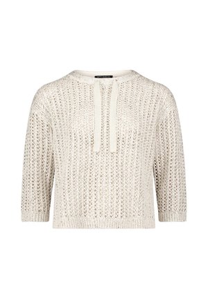 Femme aux cheveux raides et foncés portant un pull en crochet crème avec deux liens à l'avant, debout devant un fond clair uni.