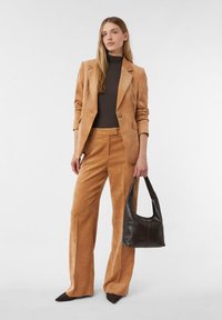 Kobieta ubrana w beżową sztruksową marynarkę i spodnie z czarnym golfem, trzymająca czarną skórzaną torebkę na tle jednolitego jasnego tła.