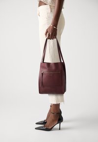 Bordeaux læder tote taske med en tekstureret overflade, har en top lynlås, forlommer og forlængede stropper. Sammen med hvide cropped bukser.
