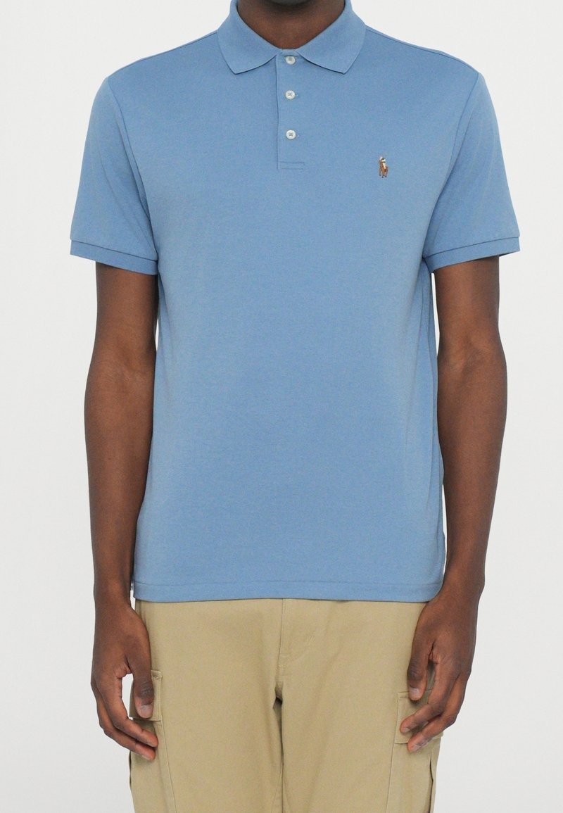 Homme portant un polo bleu clair à manches courtes avec boutons et un pantalon cargo beige, debout devant un fond uni.