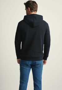 Mężczyzna stojący tyłem, ubrany w czarną bluzę z kapturem i niebieskie dżinsy na jednolitym jasnym tle.