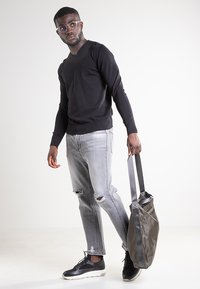 Schwarzer V-Ausschnitt-Pullover, hellgraue Jeans mit Used-Effekt, graue Sportschuhe und eine olivgrüne Tasche mit schwarzen Trägern. Lässiger, moderner Look.