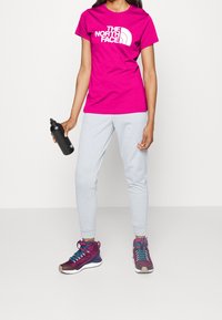 Camiseta de algodón magenta con logo blanco, pantalones deportivos gris claro y zapatos de senderismo morados con detalles en azul. Botella de agua negra en la mano.