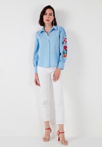 Light blue long-sleeve shirt featuring floral embroidery on sleeves, button front, and relaxed fit, paired with white trousers and multi-colored heels.