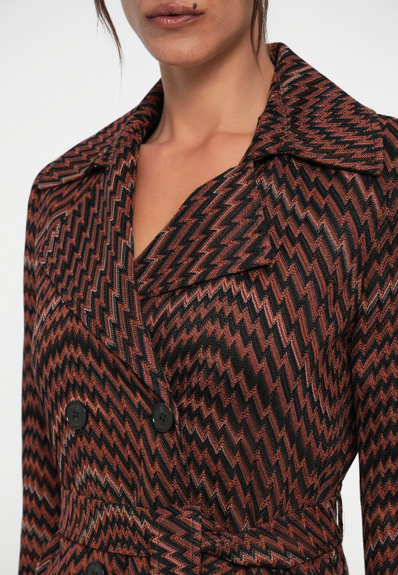 Femme portant un manteau croisé avec un motif zigzag dans des nuances de marron, noir et rose, avec un large col et une taille ceinturée.
