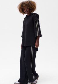 Zwarte hoodie met brede mouwen, voorzien van getextureerde zilveren strepen. Gemaakt van zachte stof en gecombineerd met zwarte wijde broek.