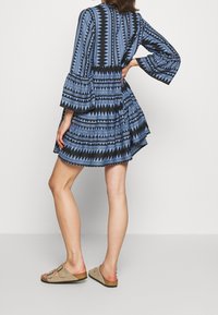 Robe bleue à motifs avec manches évasées, présentant des imprimés géométriques en noir et bleu clair. Portée avec des sandales beiges.