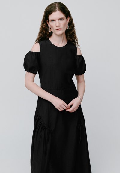 Frau mit lockigem braunem Haar, die ein schwarzes Kleid mit ausgeschnittenen Schultern und silberne Creolen trägt, steht vor einem schlichten grauen Hintergrund.