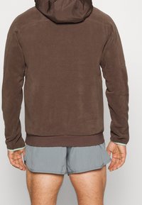 Felpa marrone con finitura testurizzata, dotata di cappuccio e sottili accenti verdi ai polsini, abbinata a pantaloni shorts grigi.