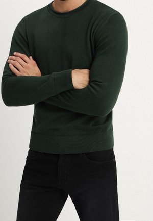 Mies, jolla on päällään tummanvihreä pyöreäkauluksinen neulepaita ja mustat housut, seisoo käsivarret ristissä vaaleaa yksiväristä taustaa vasten.