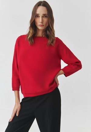 Red knitted sweater with a loose fit, round neckline, three-quarter sleeves, and textured accents along the shoulders and sleeves.