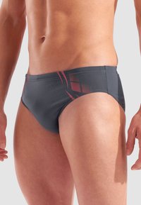 Torso maschile che indossa slip da bagno grigio scuro con linee grafiche rosse, in piedi su uno sfondo grigio chiaro semplice.