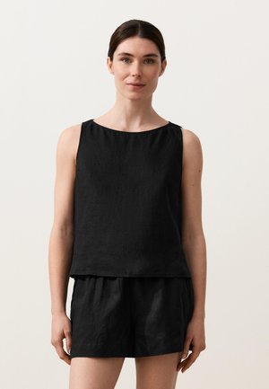 Frau mit dunklen Haaren, die ein ärmelloses schwarzes Oberteil und schwarze Shorts trägt, vor einem schlichten hellen Hintergrund stehend.