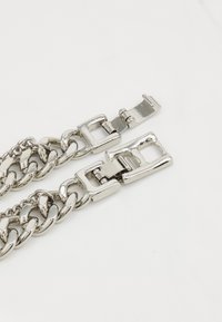 Brățară din metal argintiu, cu zale interconectate și un închizător rectangular. Textură netedă și lucioasă, cu un finisaj uniform.