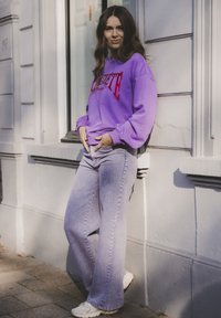 Paarse sweater met rode letters, gecombineerd met lichte denim flared jeans. Witte sneakers maken de casual outfit compleet. Lichte textuur zichtbaar.