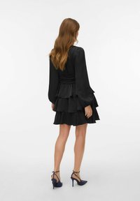 Juoda sluoksniuota suknelė su ilgomis rankovėmis; minkšta medžiaga; aptemptas bodis su raukiniu detalėmis; derinama su tamsiai mėlynais aukštakulniais batais.