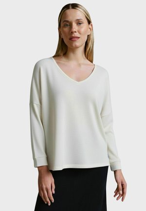 Weißer V-Ausschnitt-Pullover aus strukturiertem Stoff mit langen Ärmeln, lockerer Passform und dezenten Saumdetails. Schwarzer Rock darunter.