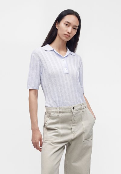 Jeune femme aux longs cheveux foncés porte un polo blanc en maille à manches courtes et un pantalon beige clair taille haute, les mains dans les poches.