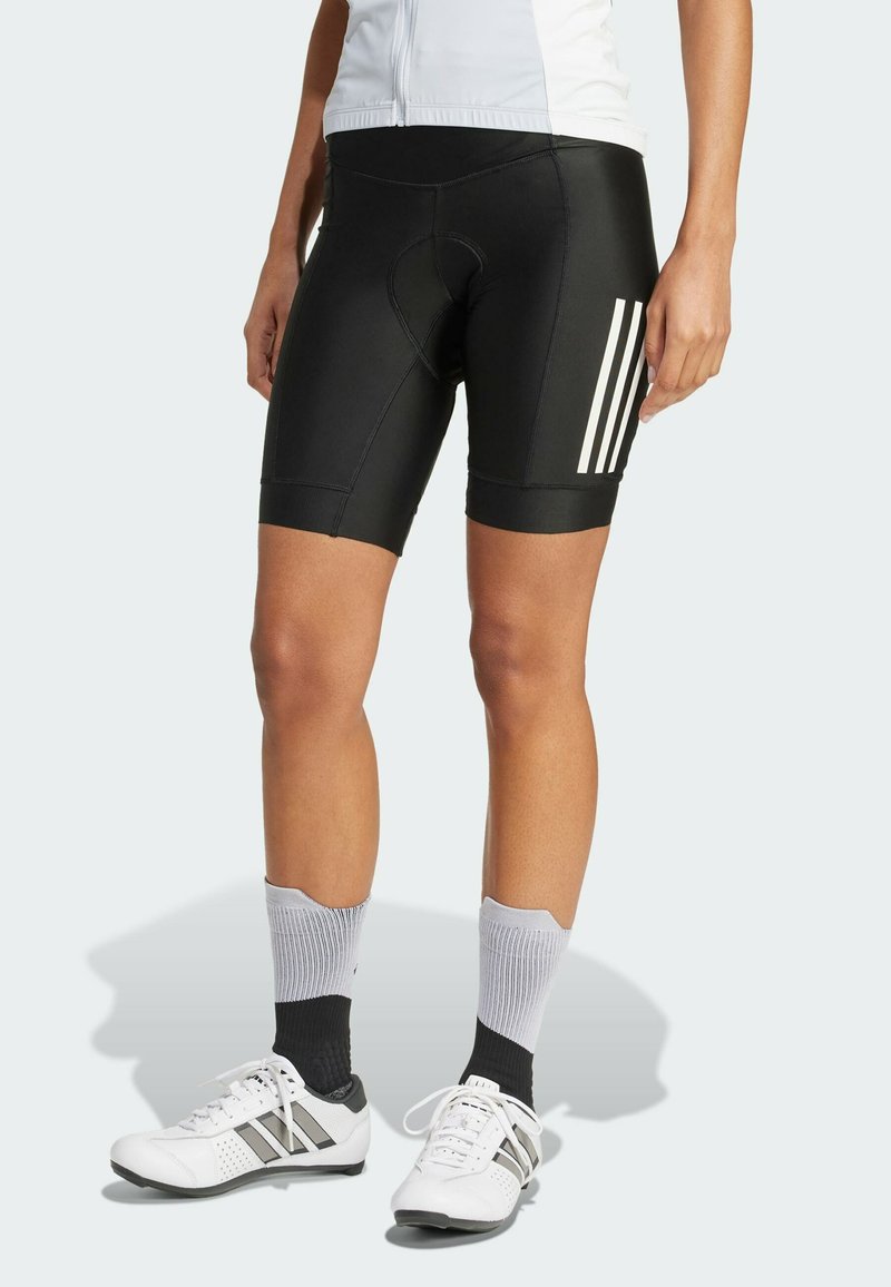 Black cycling shorts with a padded seat, featuring three white vertical stripes and a snug fit. Paired with white athletic shoes and gray socks.