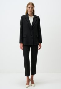 Completo nero a righe composto da un blazer con due tasche frontali e un paio di pantaloni slim-fit. Il modello indossa una maglietta bianca e scarpe col tacco a punta.