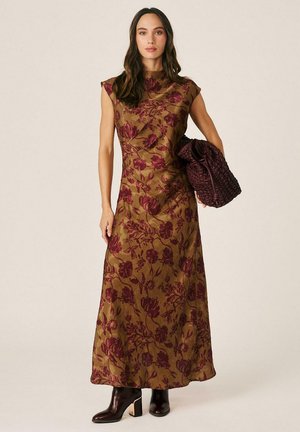 Mujer con vestido marrón sin mangas con motivos florales rojo oscuro, sosteniendo un gran bolso tejido rojo oscuro y llevando botas oscuras hasta el tobillo.