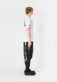Camiseta blanca de manga corta combinada con pantalones cargo negros que cuentan con múltiples bolsillos, correas y zapatos con suela texturizada. Diseño simple y funcional.