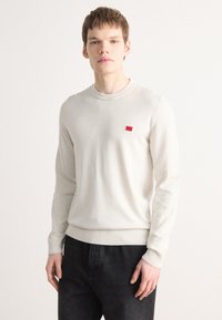 Pull crème clair à manches longues, avec un col rond et un ourlet côtelé. Petite étiquette logo rouge sur la poitrine. Associé à un jean foncé.