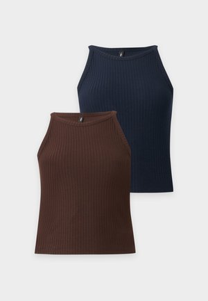 Jeune femme avec un haut marron sans manches, un jean skinny bleu avec des ourlets effilochés, des sandales à talons noires et un bracelet, posant devant un fond uni.