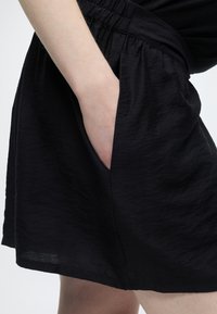 Το χέρι ενός ατόμου έχει τοποθετηθεί στην πλαϊνή τσέπη μαύρων ελαστικών σορτς με υφασμάτινη υφή.