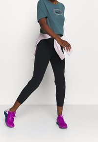 Una persona atletica che cammina indossando leggings neri, una maglietta Nike teal a maniche corte, una giacca rosa annodata in vita e scarpe da corsa viola brillante.