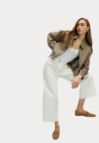Veste courte marron sur un débardeur blanc, associée à un jean blanc à jambes larges et des chaussures à enfiler marron, le tout sur fond neutre.