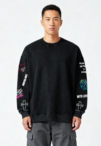 Črna majica s krožnim izrezom in grafičnimi motivi na obeh rokavih, vključno s srci, živalmi in besedilom. Izdelana iz teksturirane tkanine.