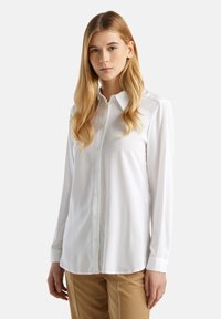 Bílá, dlouhá košile s dlouhými rukávy, vyrobená z hladkého materiálu, s klasickým límečkem a jemnými šitými detaily. V páru s béžovými kalhotami.