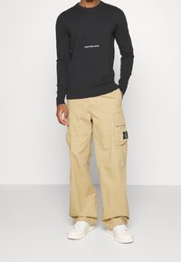 Svart crewneck-tröja med vitt "Calvin Klein Jeans"-logo, kombinerad med beige cargobyxor med sidofickor och en svart CK-lapp. Vita sneakers.