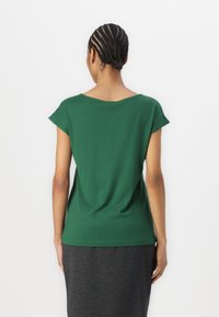 T-shirt verde a maniche corte realizzato in tessuto morbido, con una vestibilità rilassata e un ampio scollo, abbinato a una gonna grigia, vista posteriore.