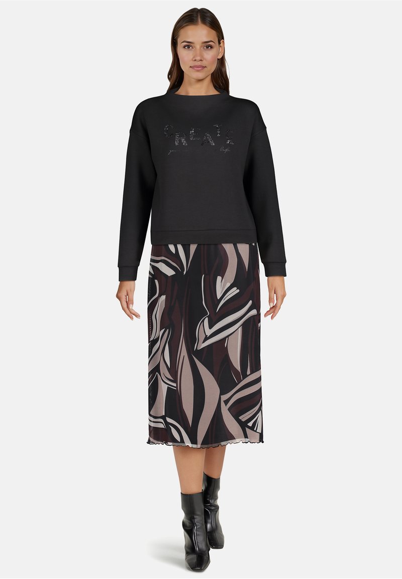 Zwarte sweatshirt met gestructureerde "Create" opdruk, gecombineerd met een midi-jurk met abstracte bladzijdenpatronen in donkere en lichte tinten. Zwarte enkellaarsjes.