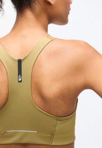 Κοντινό πλάνο του πάνω μέρους της πλάτης ενός ατόμου που φοράει αθλητικό σουτιέν racerback χακί με το λογότυπο της The North Face στο ιμάντα.