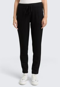 Zwarte joggerbroek van een gestructureerde stof, met een elastische tailleband en trekkoord, en tapered pijpen. G gedragen met witte sneakers.