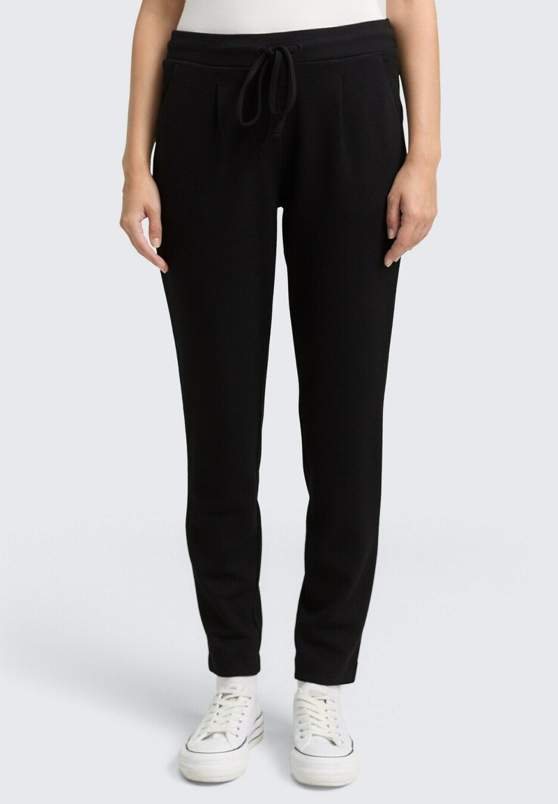 Zwarte joggerbroek van een gestructureerde stof, met een elastische tailleband en trekkoord, en tapered pijpen. G gedragen met witte sneakers.